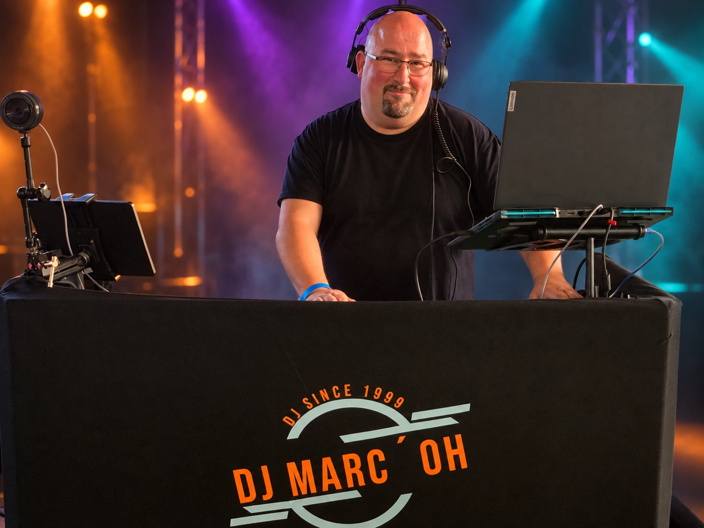 Marco Brauer alias DJ Marc’Oh an seinem DJ-Pult mit schwarzem T-Shirt vor dunklem Hintergrund mit orangefarbener, violetter und türkiser Beleuchtung